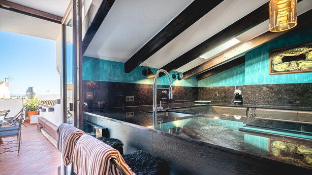 A modern kitchen with a large island, a built-in oven, and a sink. There is a window overlooking a terrace with outdoor seating.