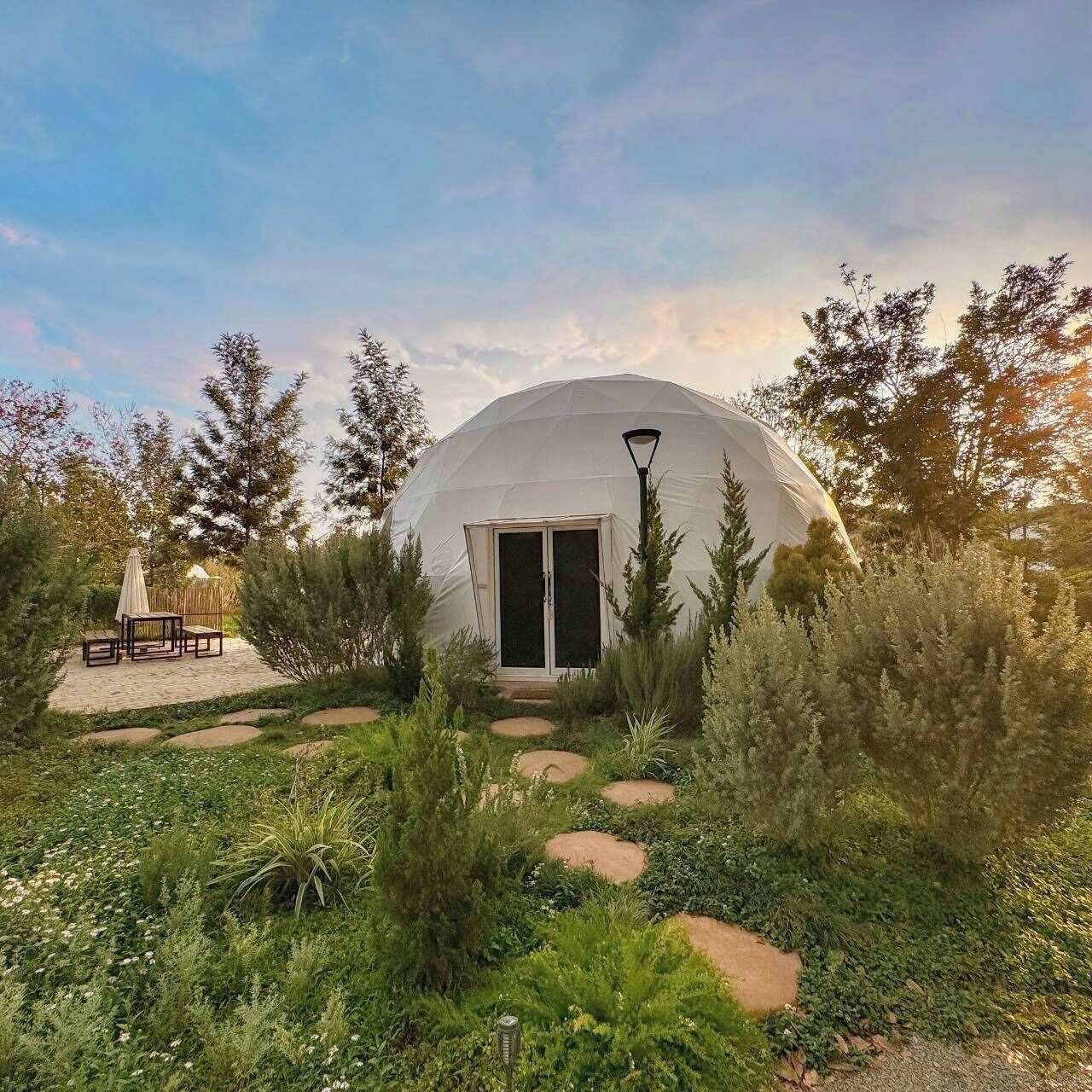 A geodesic dome structure with a door and windows, surrounded by a garden with various plants and a seating area with a table and chairs.