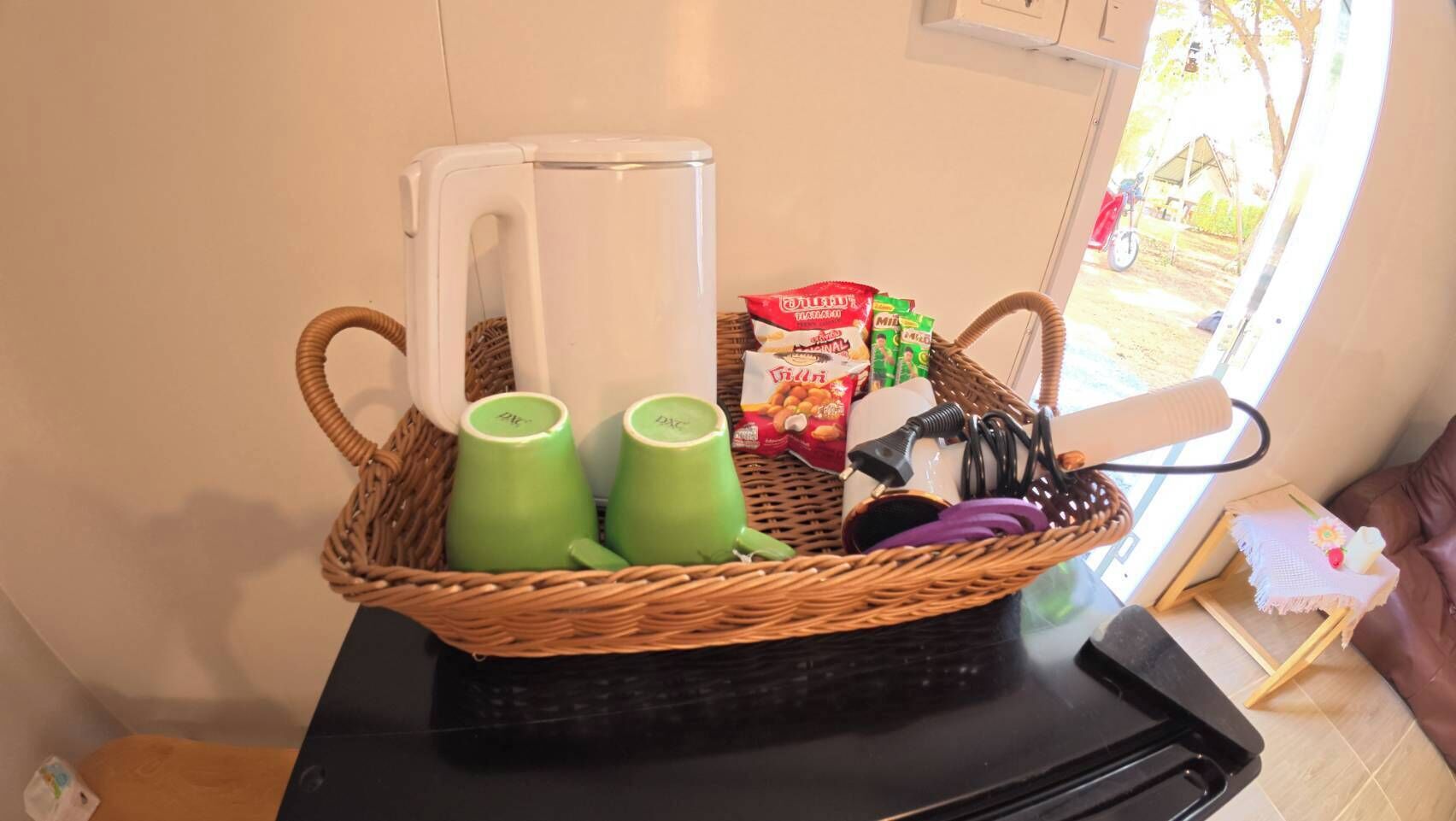 A wicker basket with green cups, a white mug, a box of snacks, and a small table with a laptop.