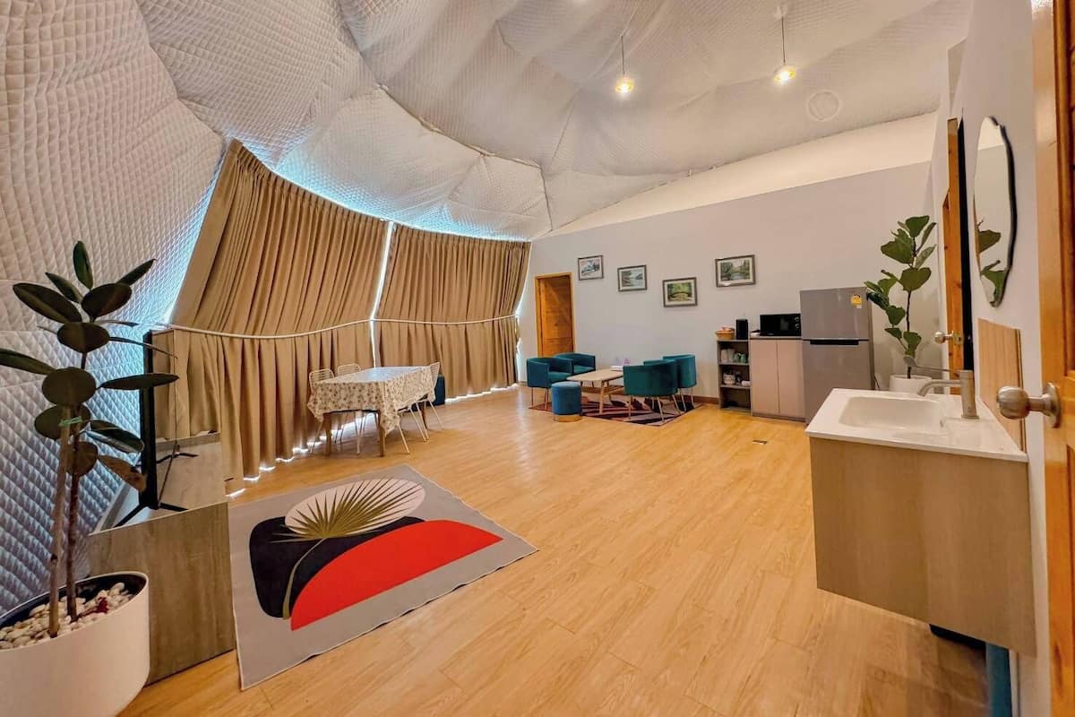 Ein modernes Wohnzimmer mit Holzboden, einem Essbereich mit Tisch und Stühlen, einer Küchenzeile mit Spüle und Schränken sowie einem großen Fenster mit Vorhängen.