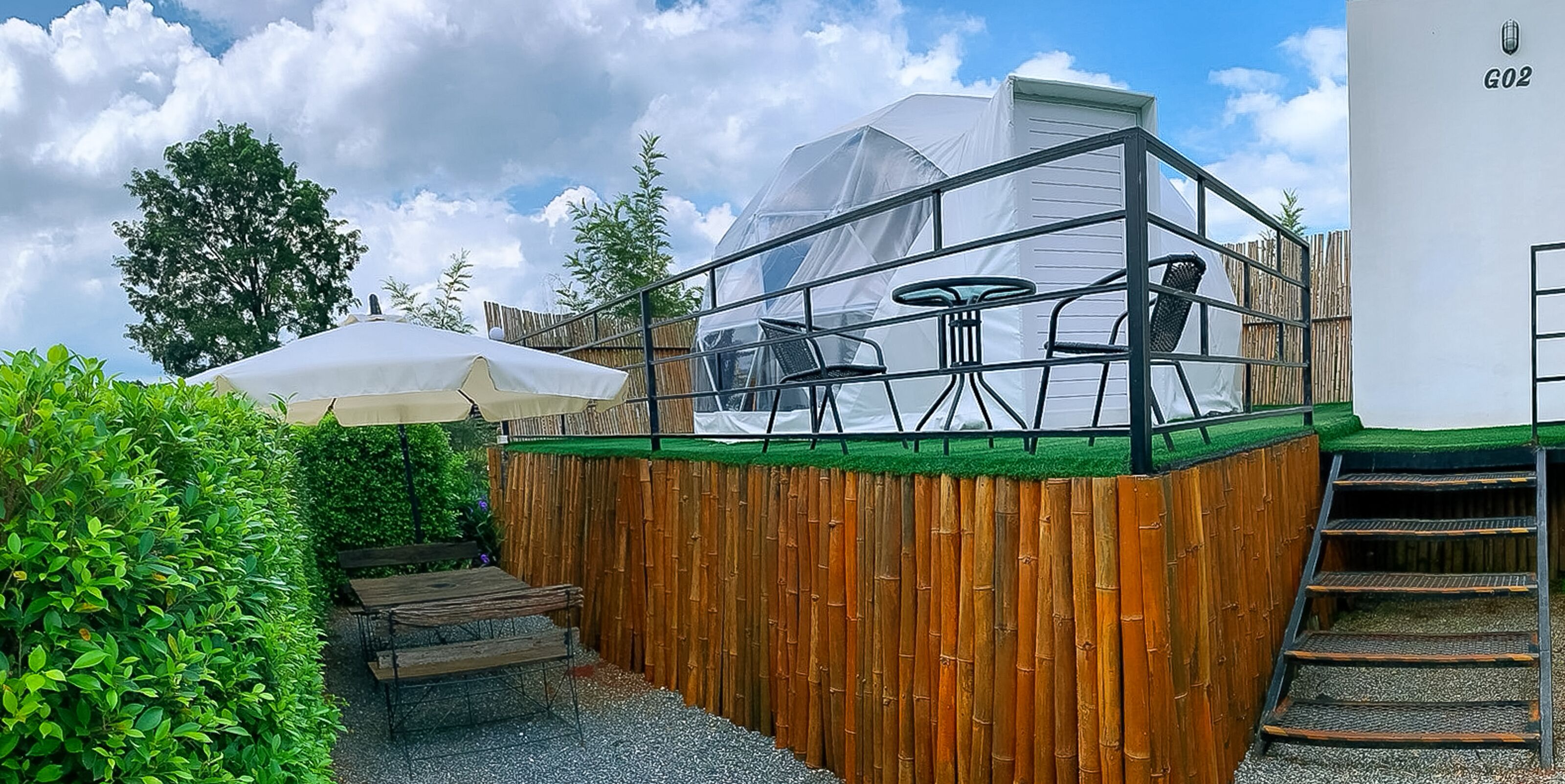 A covered outdoor seating area with a table and chairs, a wooden fence, and a white building labeled G02.