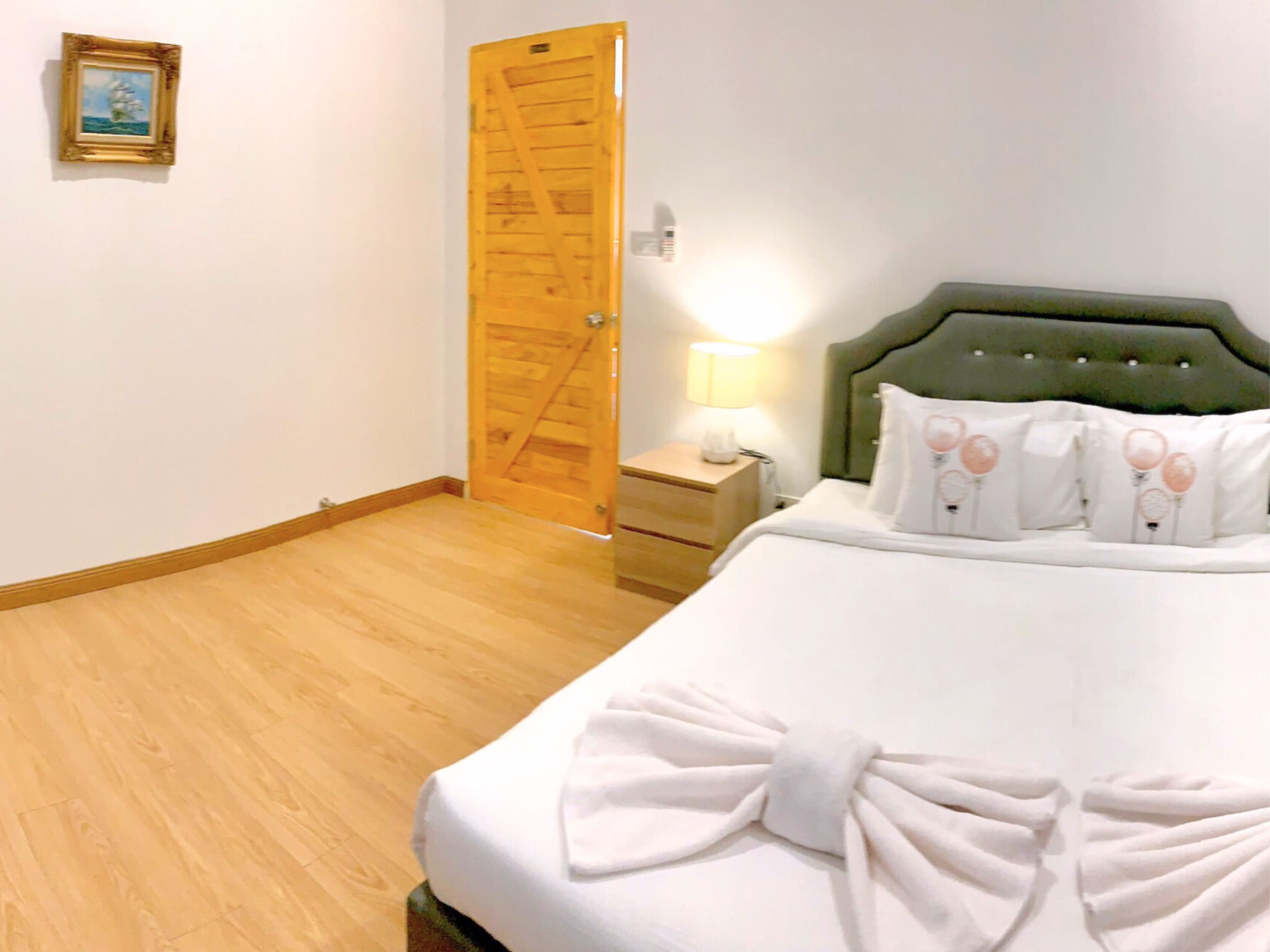 Habitación de hotel con piso de madera, cabecera tapizada de color verde, cama con ropa blanca y una toalla, mesita de noche con lámpara, y puerta de madera.