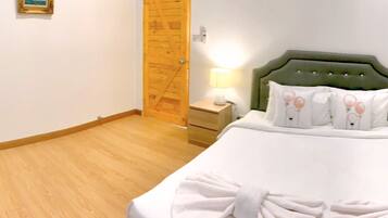 Habitación de hotel con piso de madera, cabecera tapizada de color verde, cama con ropa blanca y una toalla, mesita de noche con lámpara, y puerta de madera.