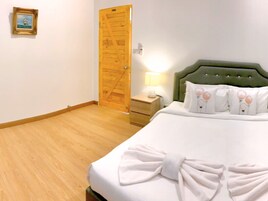 Habitación de hotel con piso de madera, cabecera tapizada de color verde, cama con ropa blanca y una toalla, mesita de noche con lámpara, y puerta de madera.