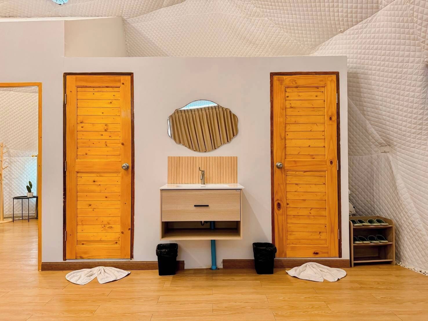 Ein Zimmer mit zwei Holztüren, einer Spiegelwand, einem Holztischchen mit Waschbecken und zwei schwarzen Mülleimern.