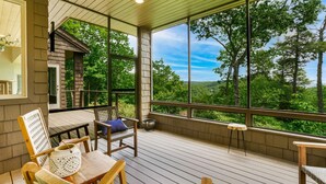 A covered outdoor deck with wooden furniture, large windows, and a view of a forested landscape. - The View From Up Here Changes Everything | Big Bear Pen (Highlands)