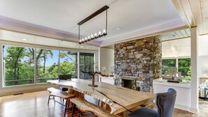A spacious dining area with a large wooden table and benches, a stone fireplace, and large windows offering a view of the outdoors. - The View From Up Here Changes Everything | Big Bear Pen (Highlands)