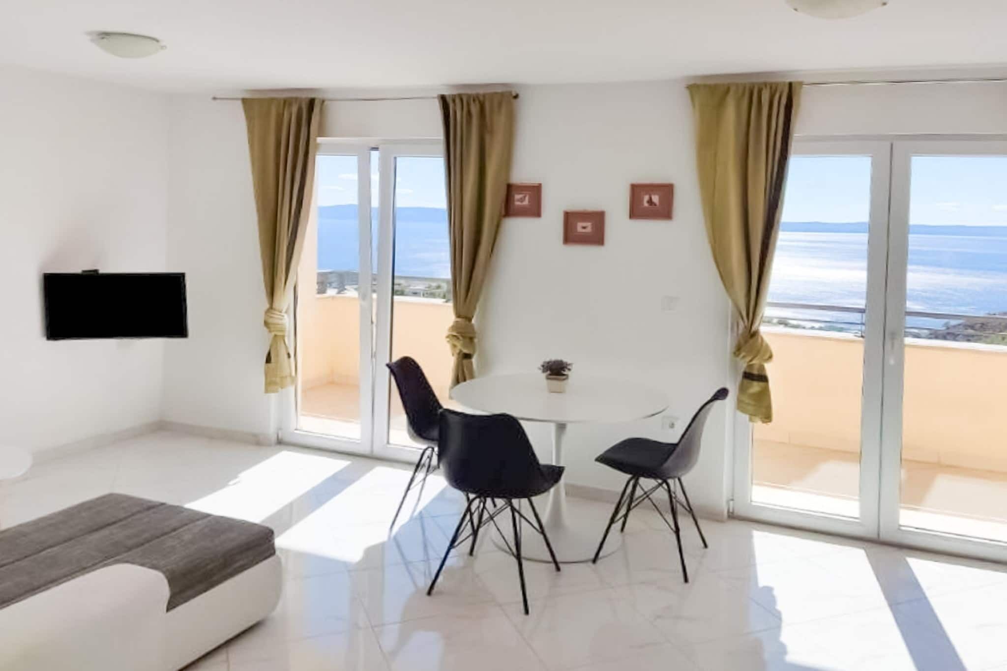 A modern living room with a sofa, a round table, and two chairs. There is a TV mounted on the wall and a view of the sea through large windows.