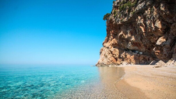A beach with clear shallow waters, a rocky cliff, and a clear sky.