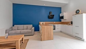 A modern living room with a blue accent wall, a grey sofa, a wooden coffee table, and a kitchen area with white cabinets and a microwave. - New, quiet, comfortable 3-star Meublé de Tourisme apartment. (Saint-Pol-de-Léon)