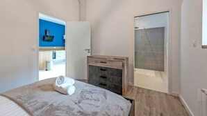 A bedroom with a bed, a wooden dresser, a bathroom with a shower, and a kitchen visible through an open door. - New, quiet, comfortable 3-star Meublé de Tourisme apartment. (Saint-Pol-de-Léon)