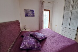A bedroom with a bed, purple bedding, a nightstand, a lamp, a painting, a window with curtains, and a wardrobe.