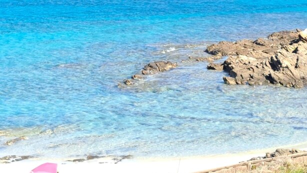 A beach with clear blue water, rocky outcrops, and a wooden fence.