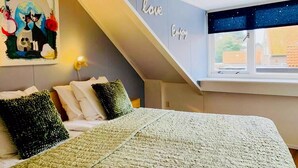 A bedroom with a sloped ceiling, a bed with a green and white blanket, a painting on the wall, and a window with a starry night view. - Nice holiday residence in De Koog near the beach (De Koog)