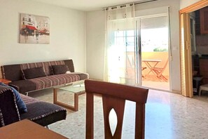 A living room with a sofa, a glass coffee table, and a wooden chair. There is a sliding glass door leading to an outdoor area with a table and chairs.