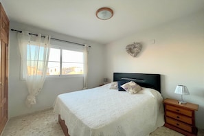 Un dormitorio con cama, mesita de noche, ventana con cortinas, un adorno en la pared y una puerta.