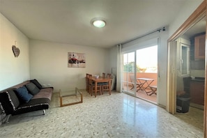 A living room with a sofa, a dining table, and chairs, a glass coffee table, a ceiling light, and a sliding glass door leading to an outdoor patio.