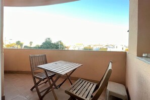 Ein Balkon mit Holztisch und zwei Stühlen, einem kleinen Hocker und Blick auf Gebäude und Bäume.