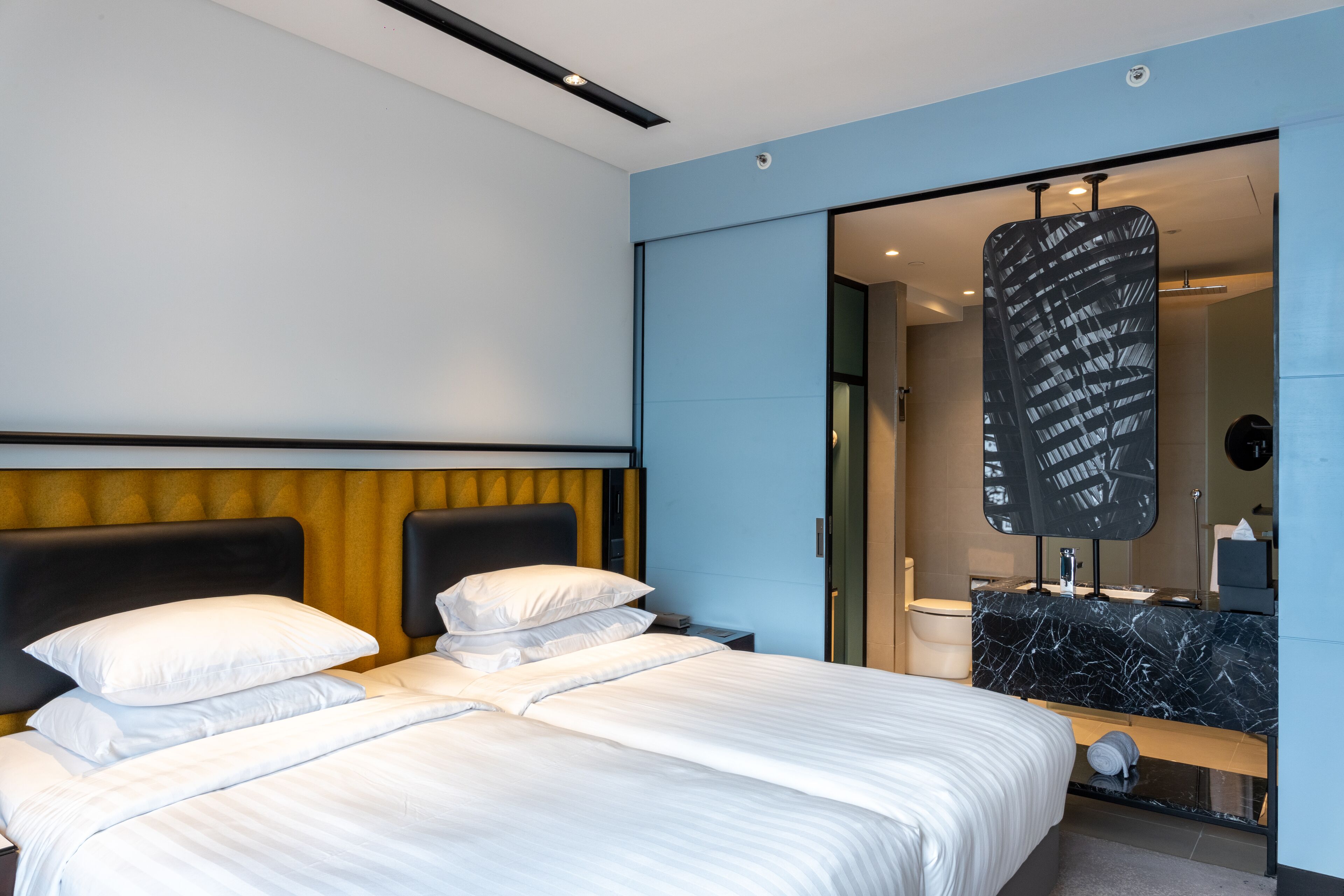 A modern hotel room with a large bed, a headboard with a yellow accent, a black marble bathroom vanity, and a large mirror.