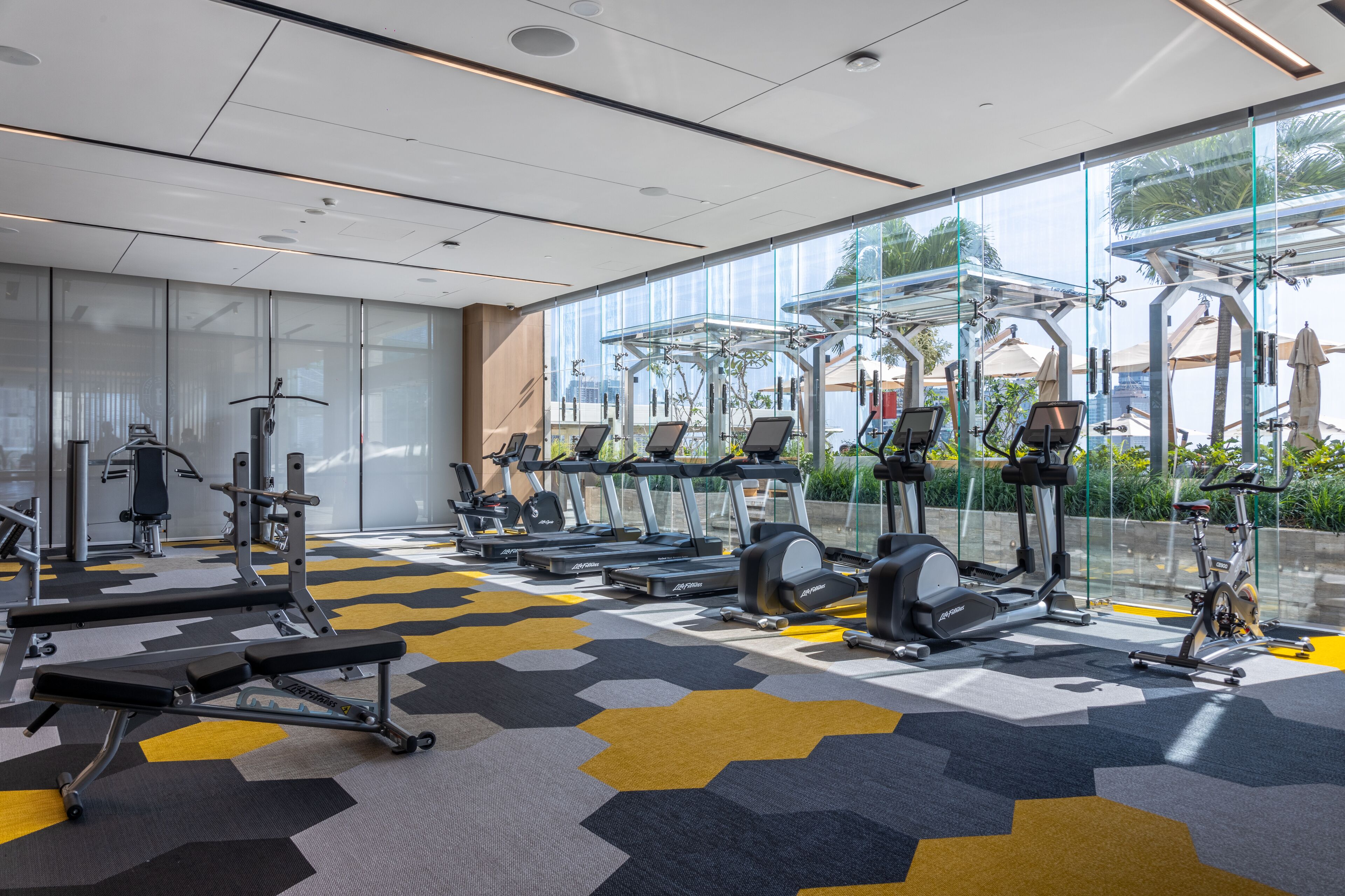 A modern gym with various exercise equipment, large windows, and a patterned floor.