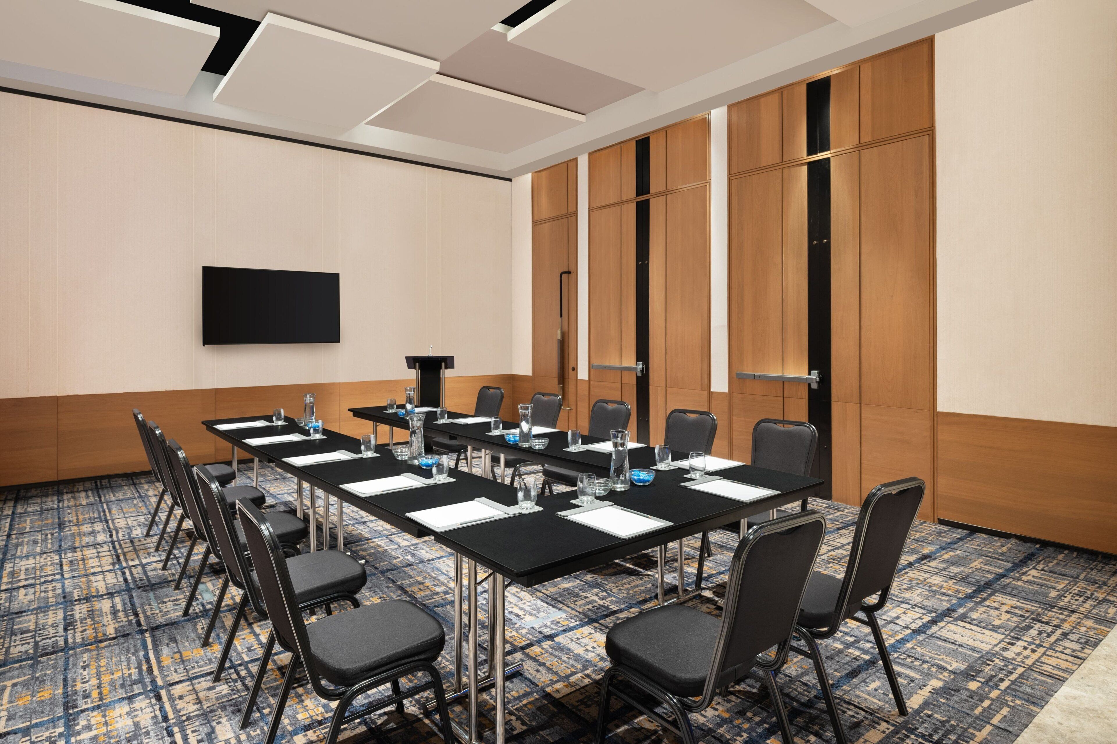 A conference room with a long table, chairs, a flat-screen TV, and a patterned carpet.
