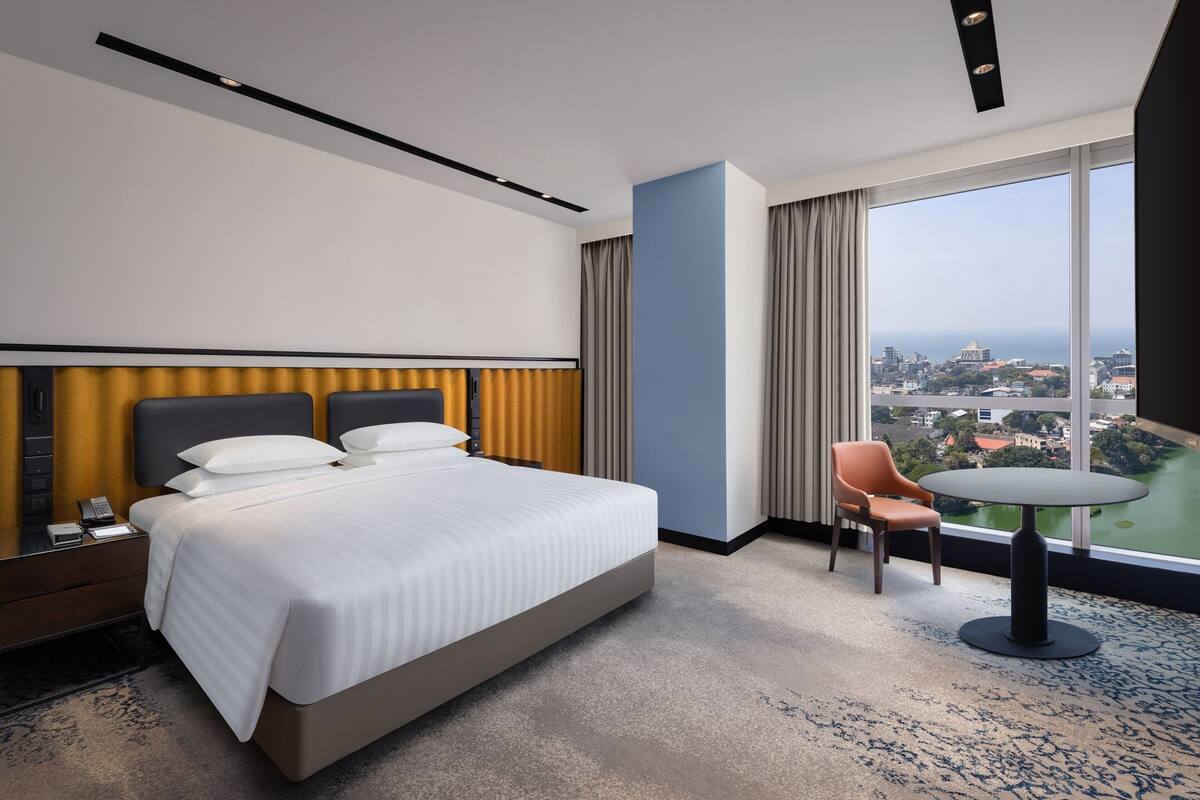 A hotel room with a large bed, a desk, a chair, and a view of a cityscape through large windows.
