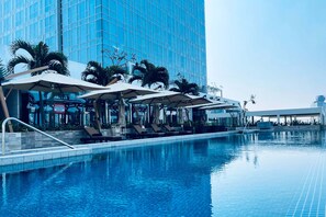 A modern hotel pool area with lounge chairs and umbrellas, surrounded by palm trees and a tall glass building.