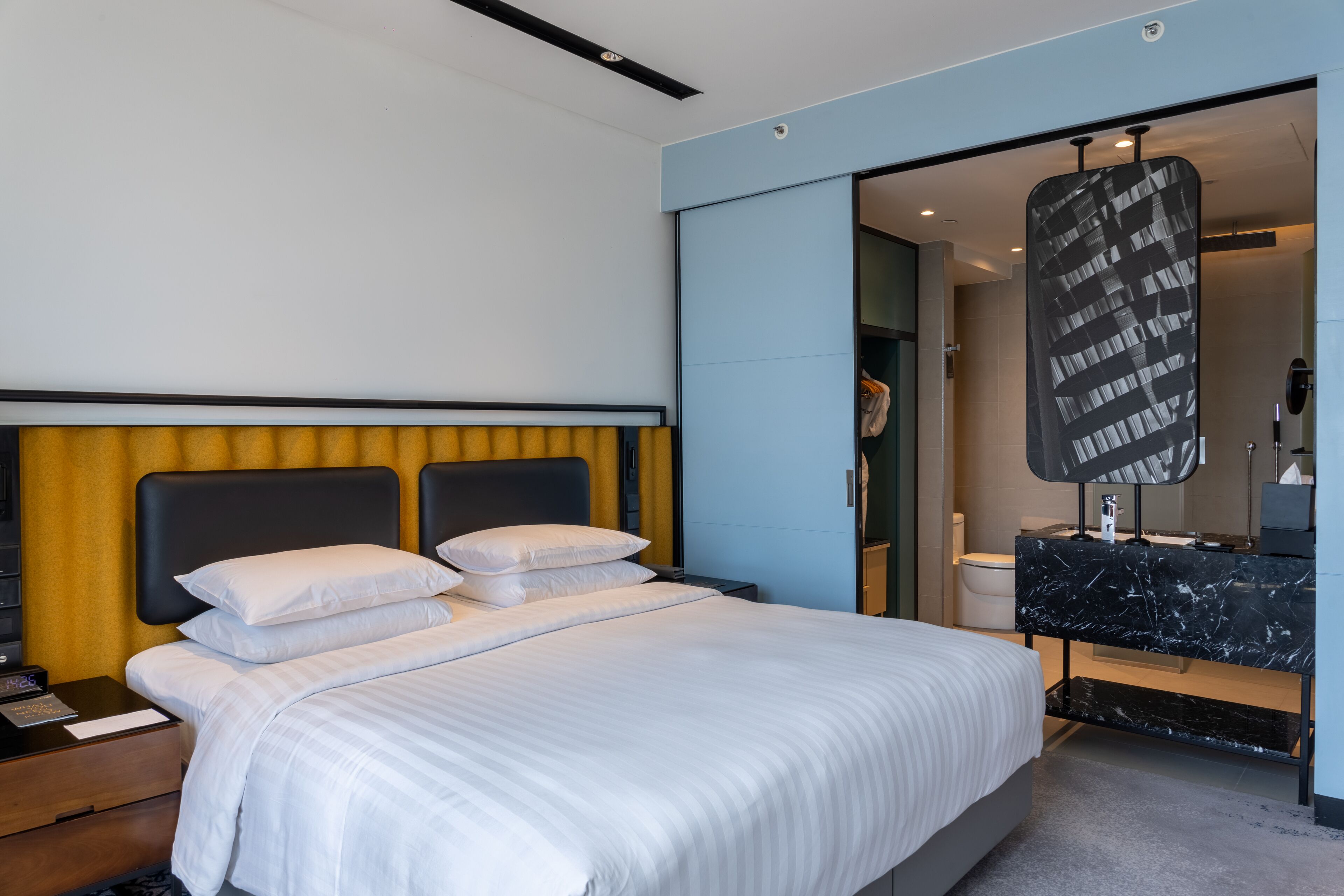 Una habitación de hotel moderna con una cama grande, una mesita de noche, un espejo y un baño visible a través de una puerta abierta.