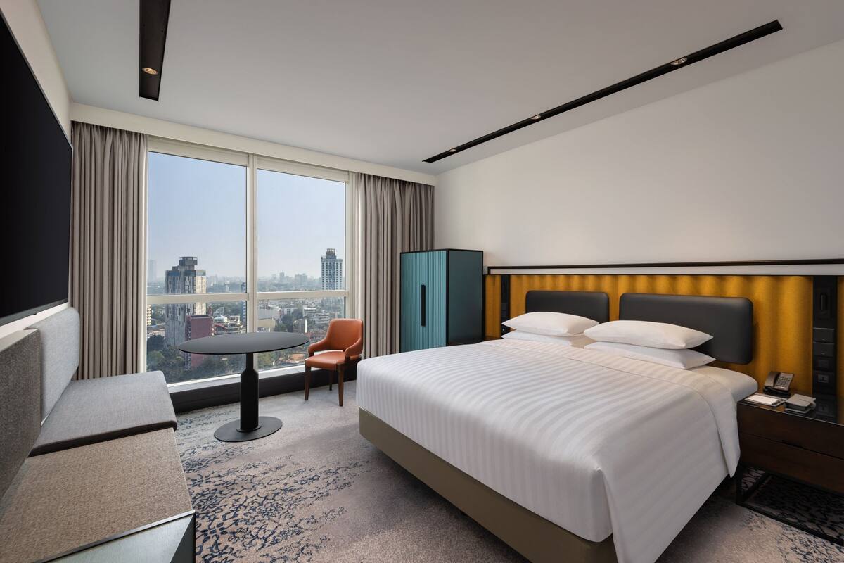 A hotel room with a large bed, a desk, a chair, a small table, and a view of the cityscape through large windows.