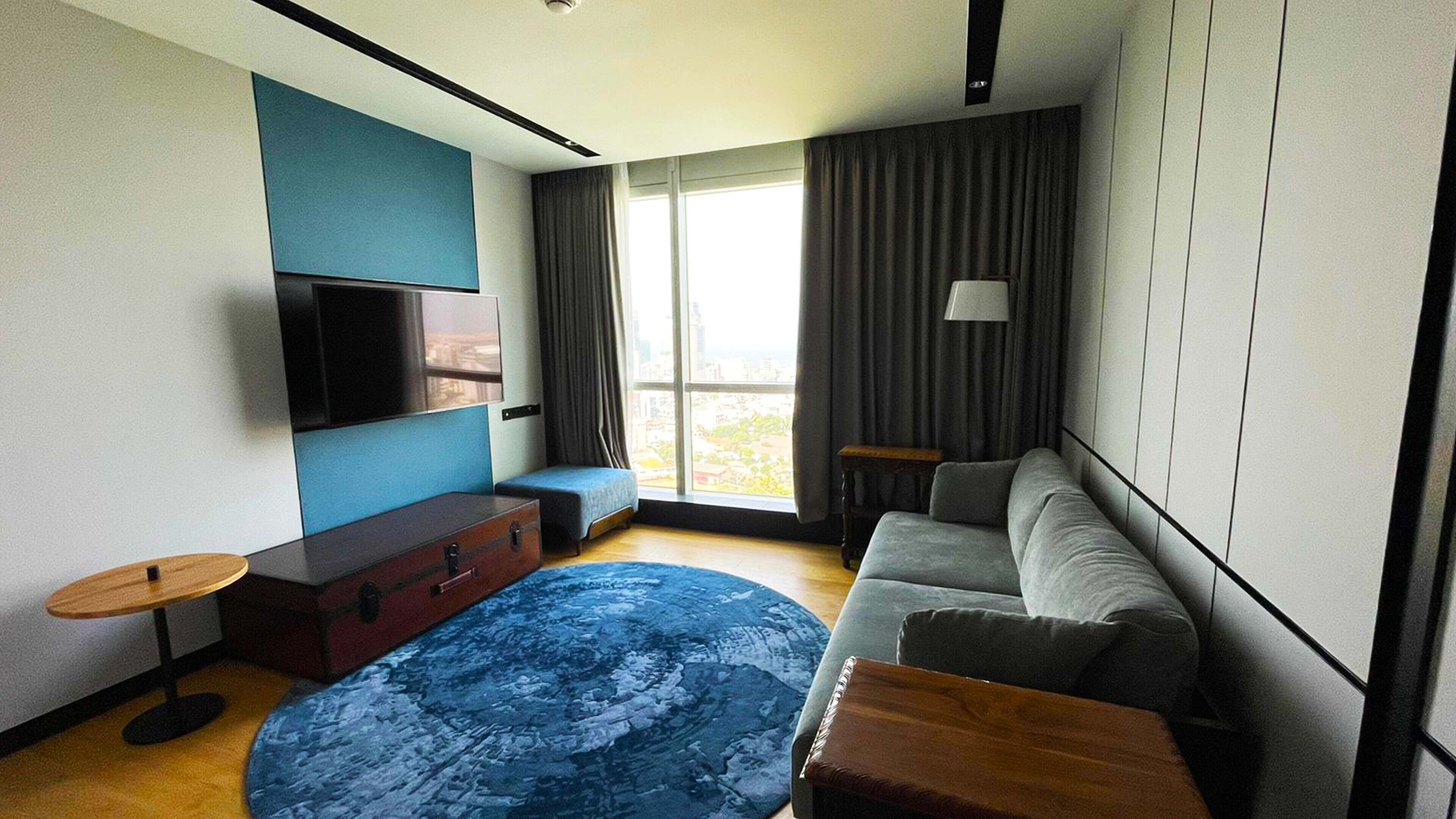 A modern living room with a sofa, a flat-screen TV, a wooden coffee table, a blue rug, and a large window with curtains.