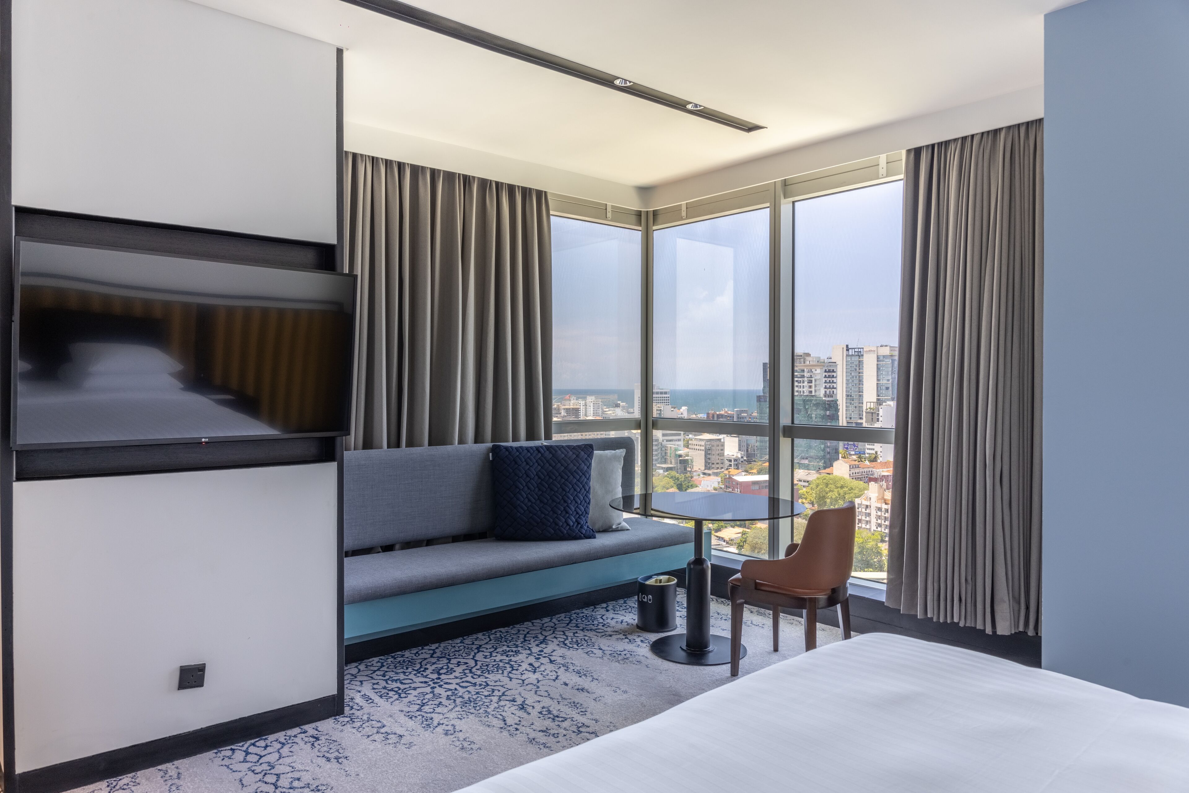 A modern hotel room with a large window, a sofa, a small table, and a bed.