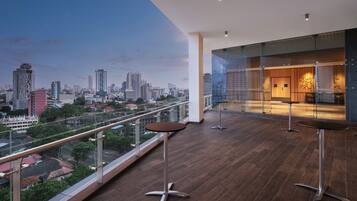 A modern hotel balcony with a city view, glass walls, and two bar stools.