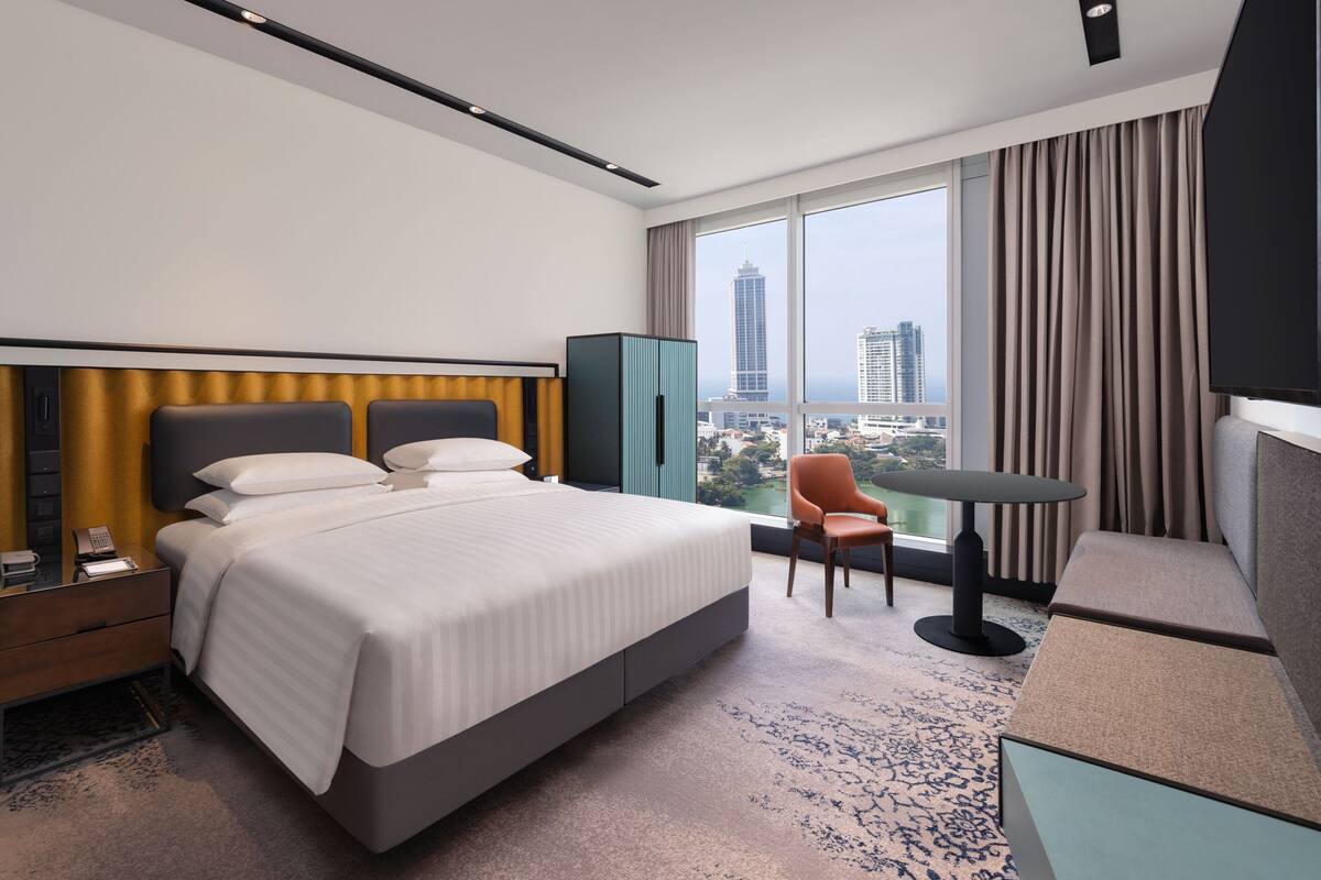 A modern hotel room with a large bed, a desk, a chair, and a view of the cityscape through large windows.