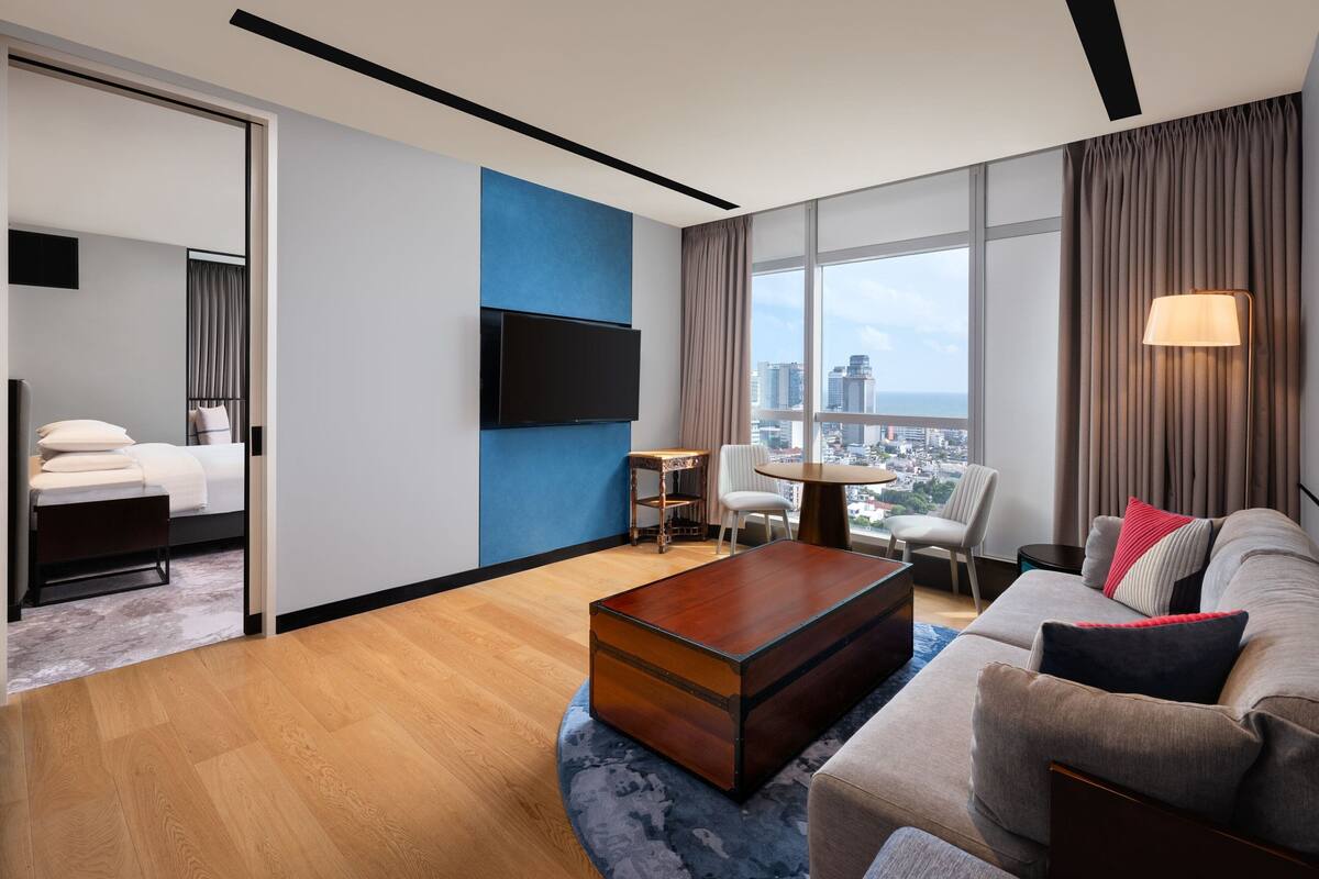 A modern hotel room with a large window, a flat-screen TV, a sofa, a coffee table, and a dining area with a table and chairs.