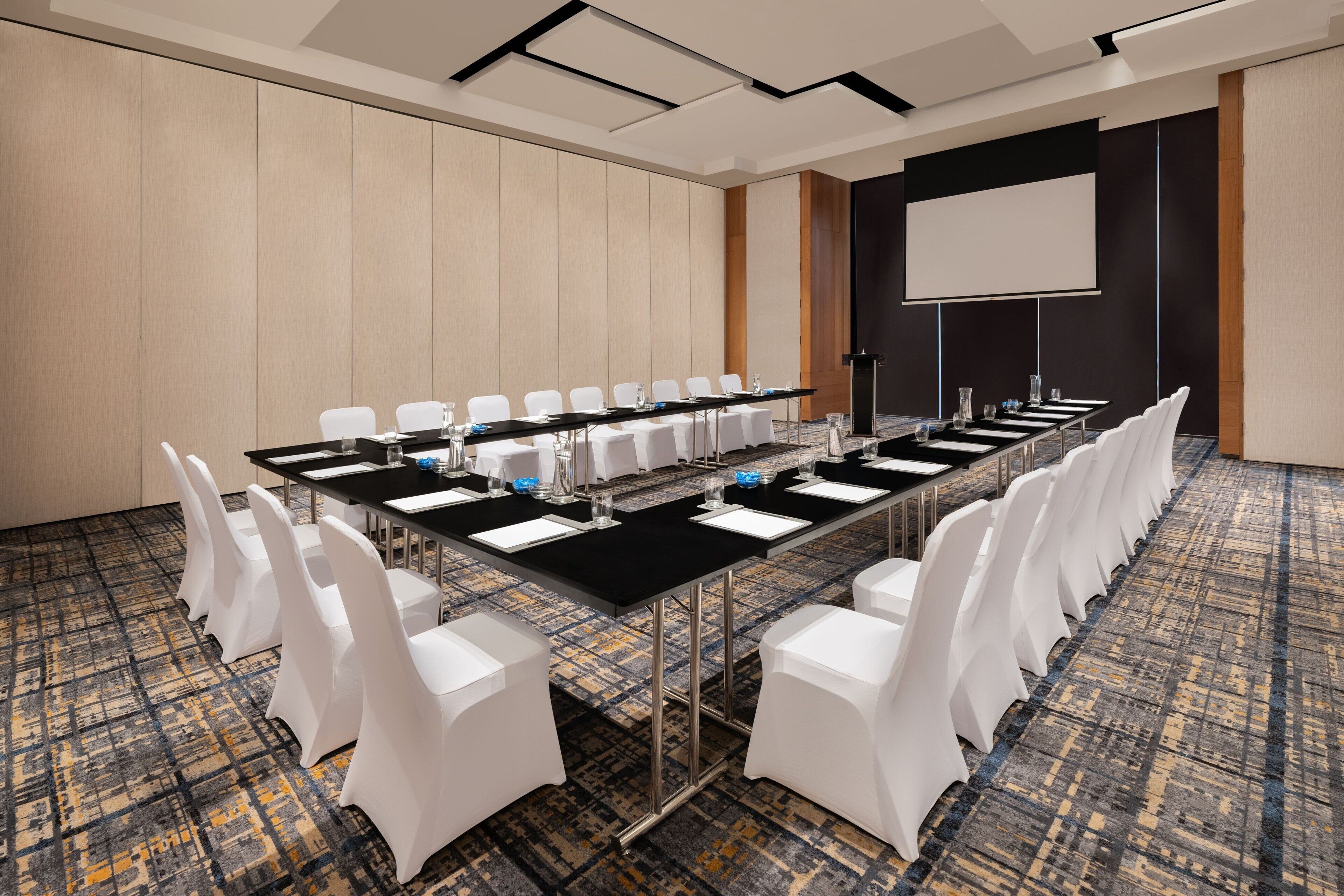 A conference room with a long table, white chairs, a projector screen, and a carpeted floor.