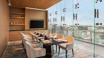 A conference room with a long table, chairs, a flat-screen TV, and a city view through large windows.