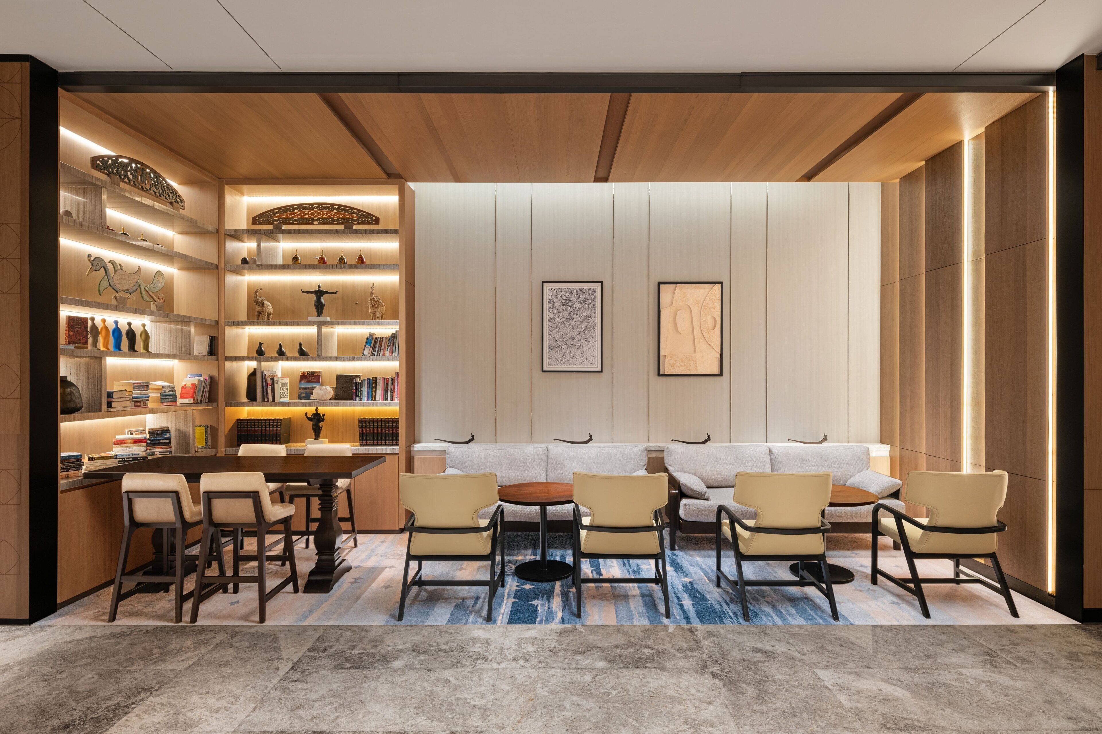 A modern interior with a wooden ceiling, a central seating area with a sofa and chairs, a dining table with chairs, and a bookshelf filled with books and decorative items.