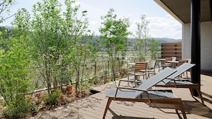 Terraza de madera con sillones de descanso y una mesa, con vistas a un jardín ajardinado y montañas distantes.