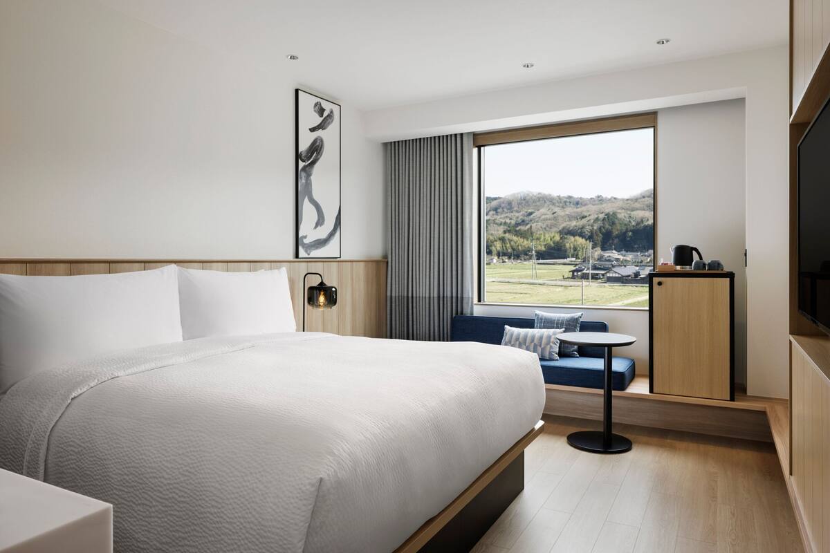 Ein modernes Hotelzimmer mit einem großen Bett, einem kleinen Tisch, einer Couch und Blick auf eine grüne Landschaft durch ein großes Fenster.
