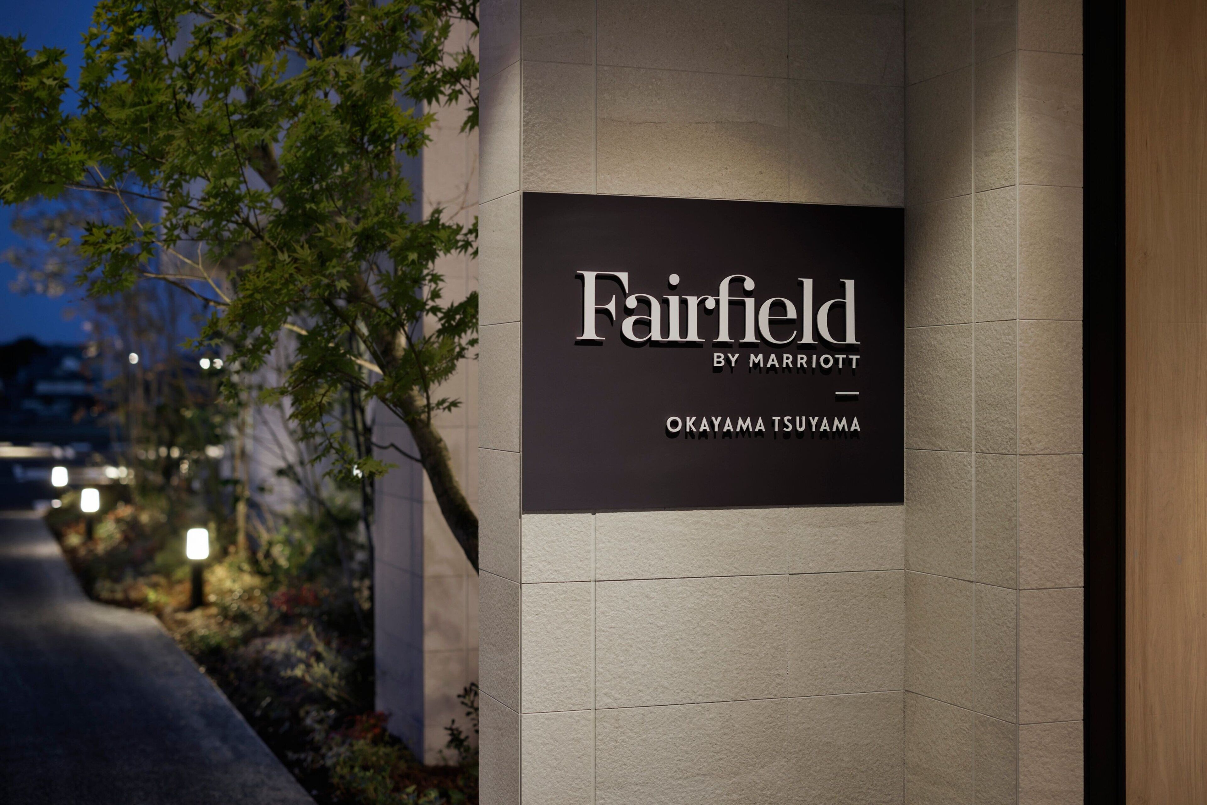 A sign for Fairfield by Marriott in a well-lit urban area with a paved walkway and greenery.