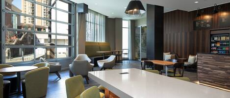 A modern interior with a long communal table, several chairs, a bar area, and large windows offering a view of the cityscape.