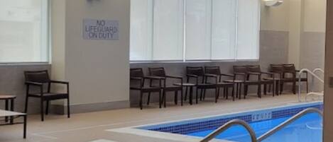 Une piscine intérieure avec un panneau indiquant « Aucun sauveteur de service », des chaises disposées le long du bord de la piscine et une vue dégagée sur le bord du bassin.