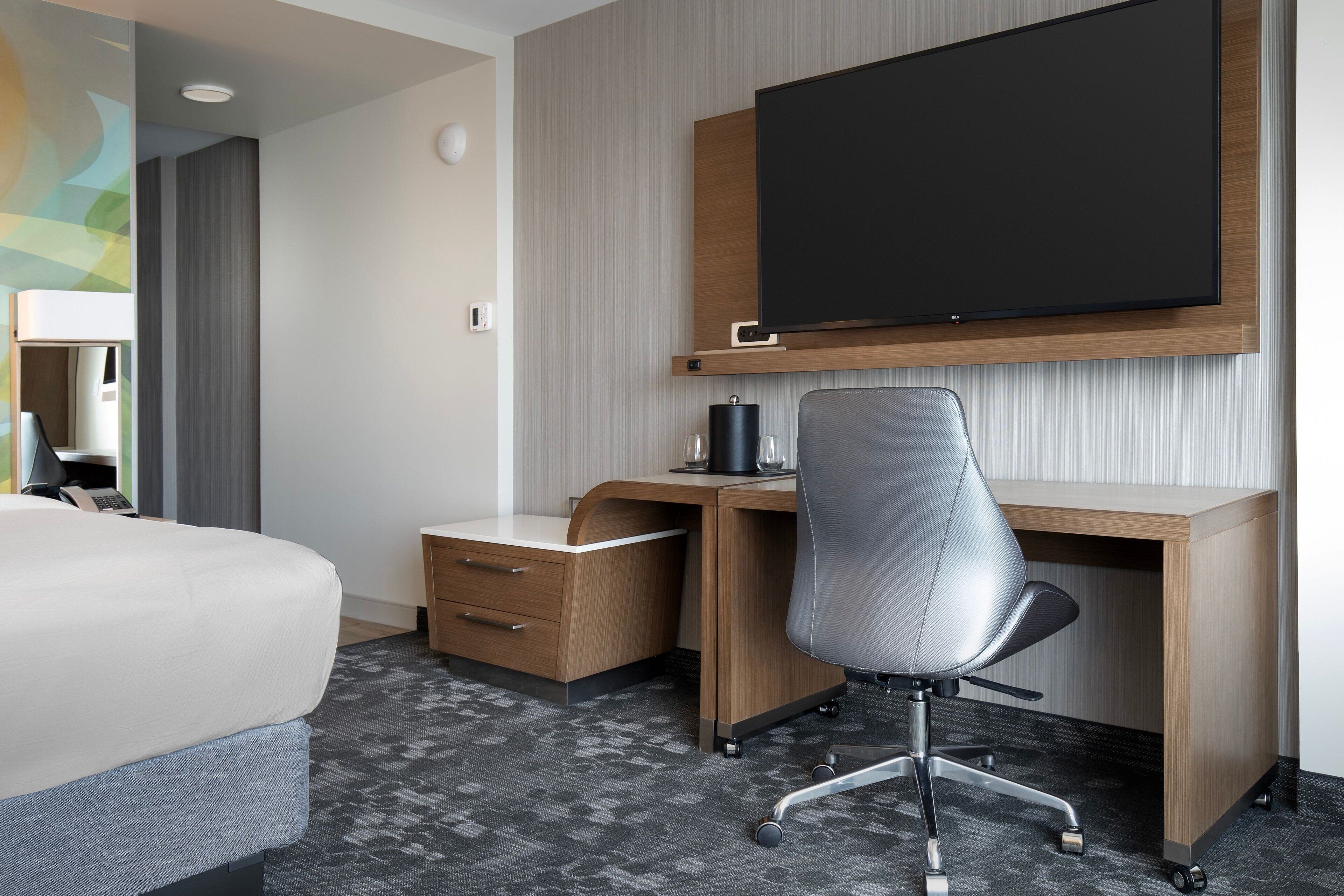 Hotellihuone, jossa on sänky, työpöytä tuolilla, taulutelevisio ja pieni laatikostokaappi.
