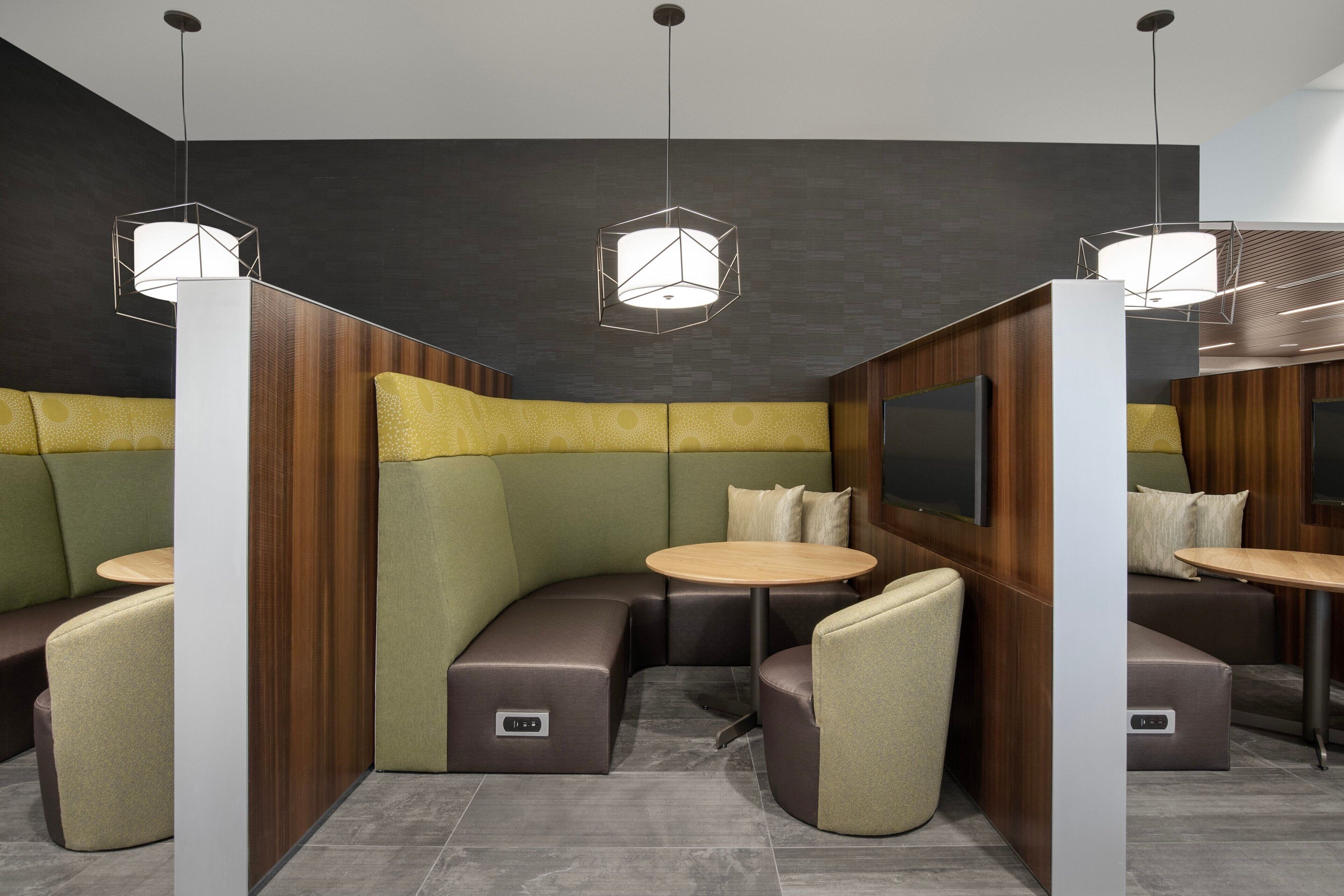 Un área de estar moderna con asientos acolchados y mesas redondas, con lámparas colgantes geométricas y una pared oscura con detalles de paneles de madera.