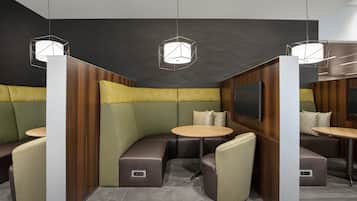 Un área de estar moderna con asientos acolchados y mesas redondas, con lámparas colgantes geométricas y una pared oscura con detalles de paneles de madera.