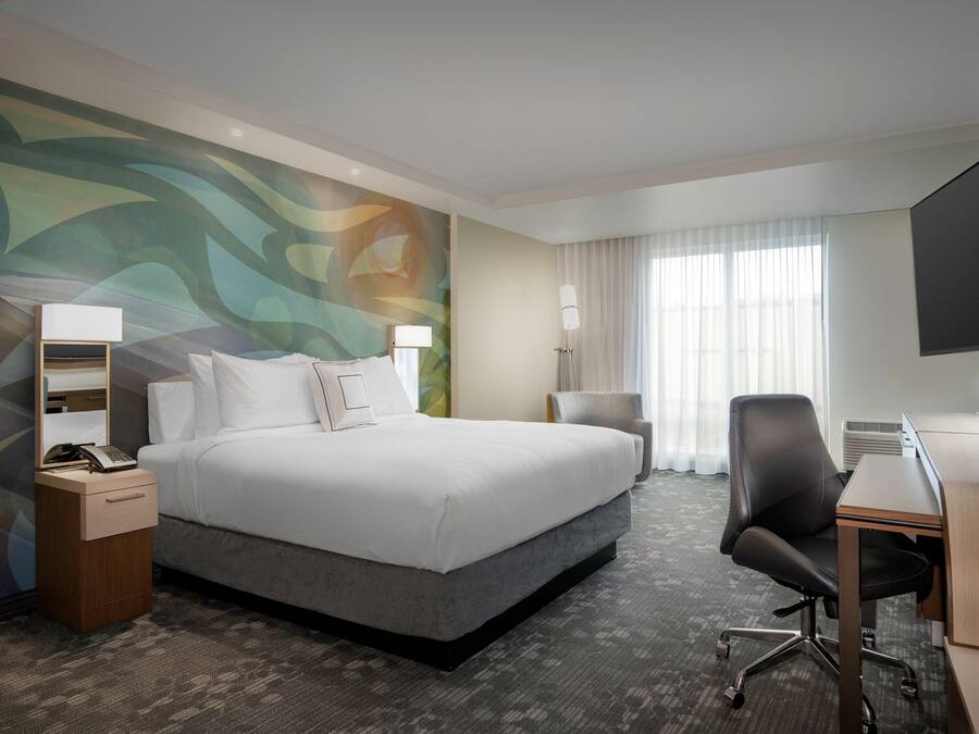 A hotel room with a large bed, a desk, a chair, a TV, a lamp, and a wall with a colorful abstract design.