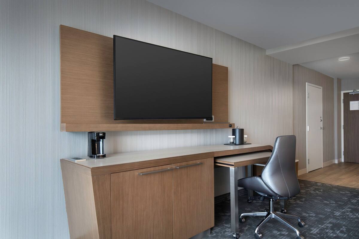 Een moderne hotelkamer met een houten bureau, een flatscreen televisie, een koffiezetapparaat en een zwarte kantoorstoel.