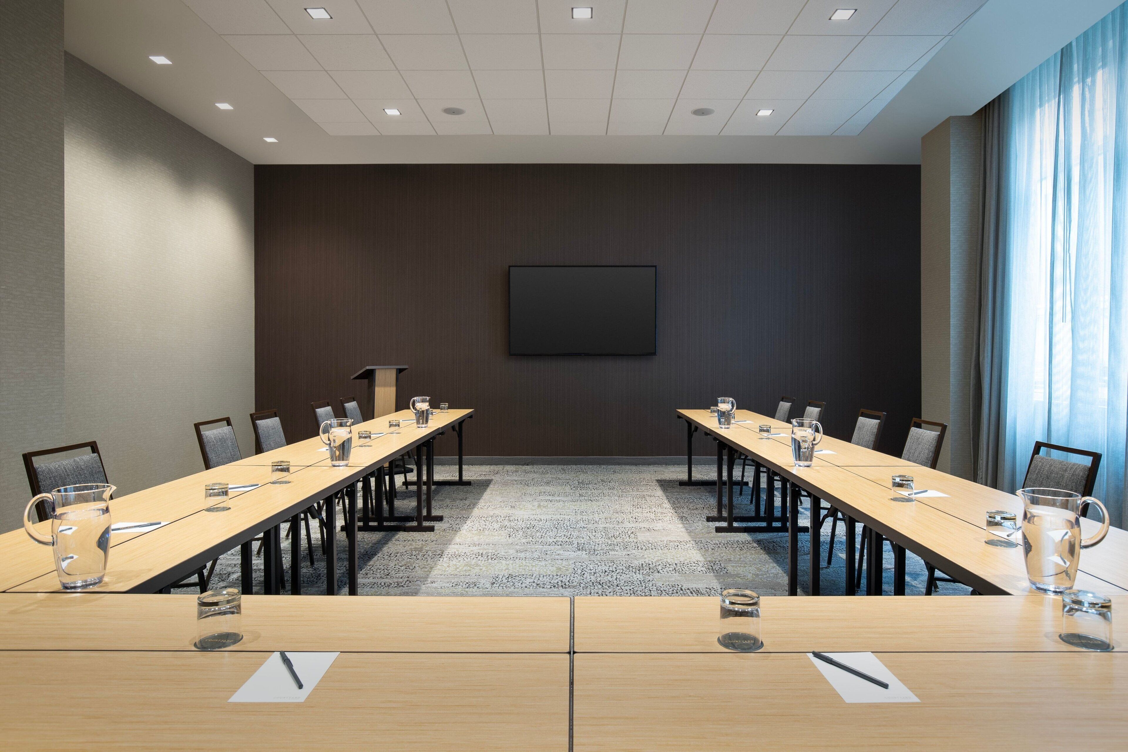 A conference room with a long table, chairs, a projector screen, and a window with curtains.