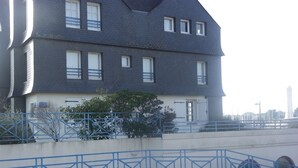 Un immeuble à plusieurs étages, doté d’un toit sombre et de murs blancs, avec une balustrade bleue et un petit arbre devant.