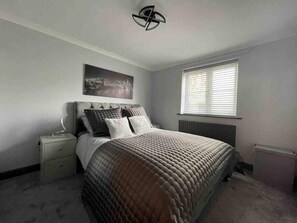 Une chambre à coucher comprenant un lit, une table de chevet, un radiateur, une fenêtre avec des stores et un luminaire fixé au mur.